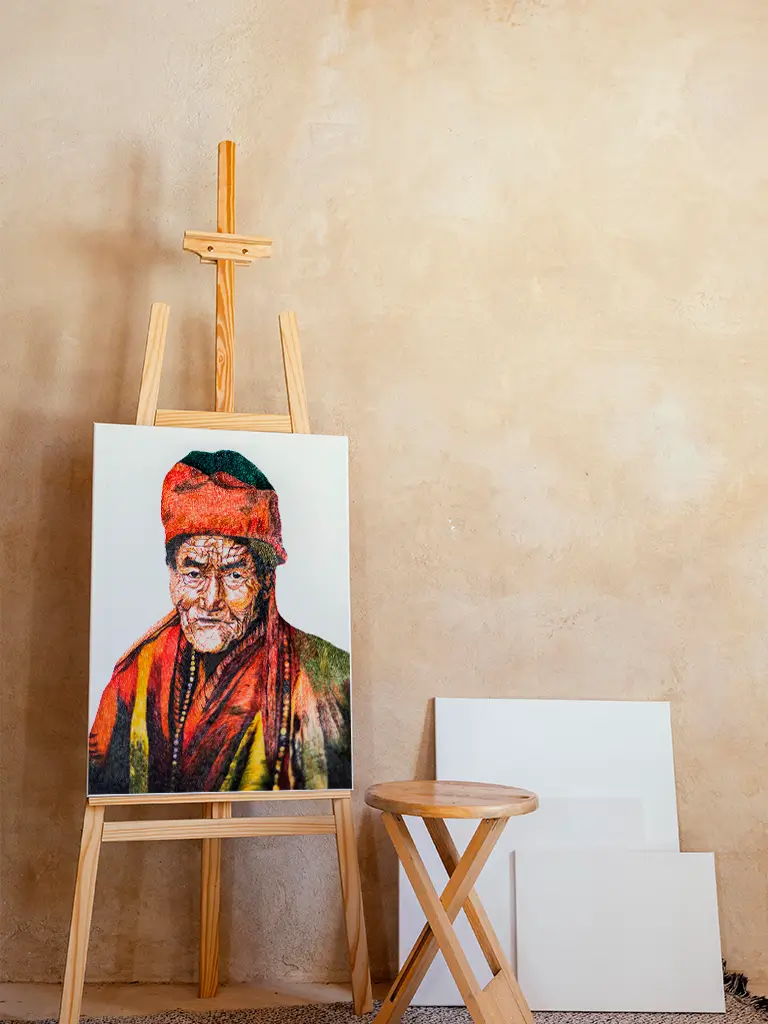 Retrato colorido de una persona anciana con vestimenta tradicional, en un caballete de madera clara junto a lienzos blancos y una banqueta