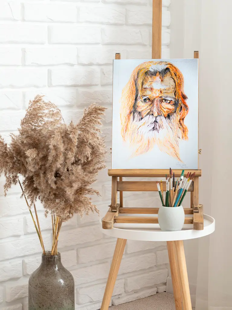 Retrato artístico de un hombre anciano con barba blanca y cabello largo en tonos cálidos, colocado en un caballete sobre una mesa blanca junto a un jarrón con pampas secas y pinceles.