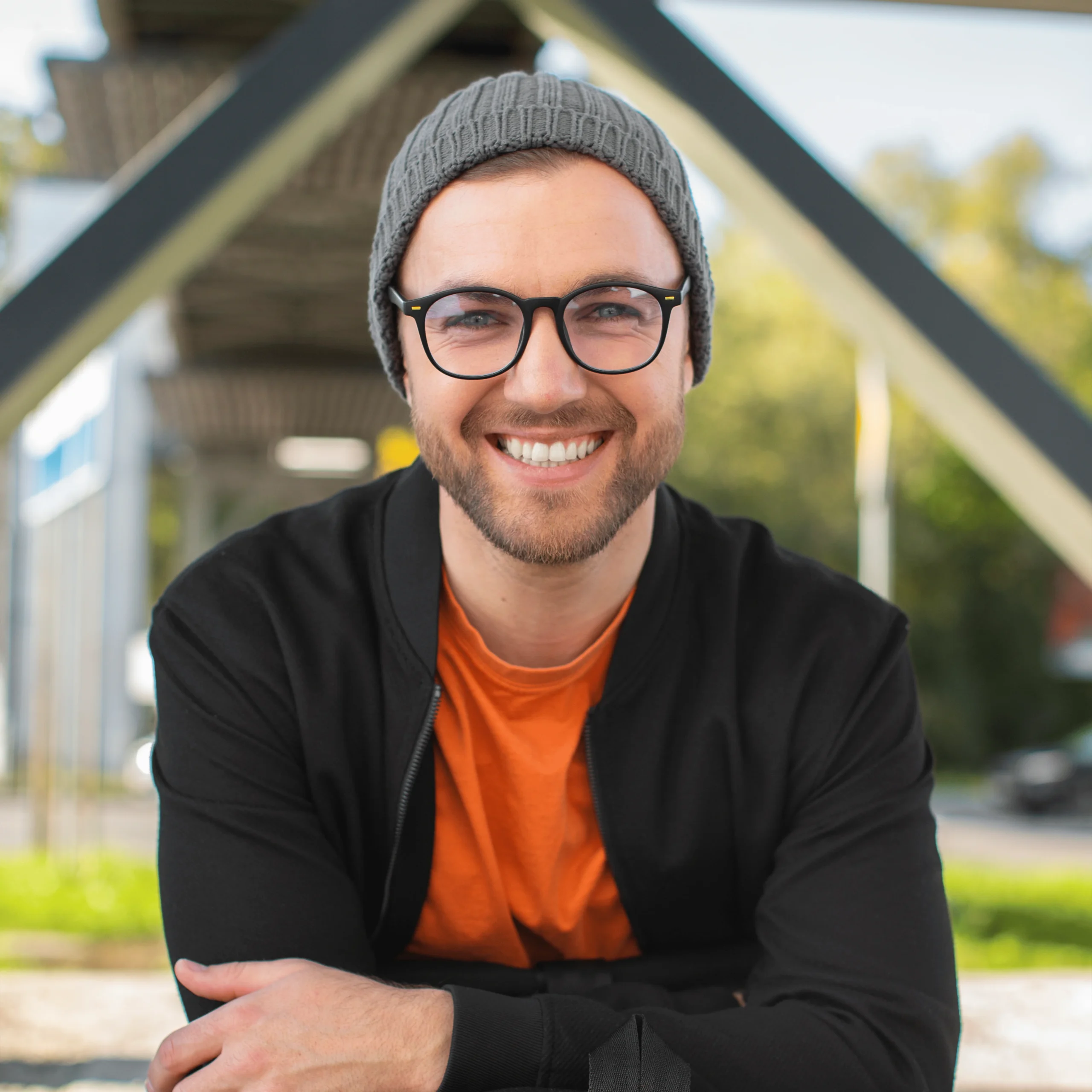 Hombre joven con gafas y gorro gris sonriendo al aire libre.
